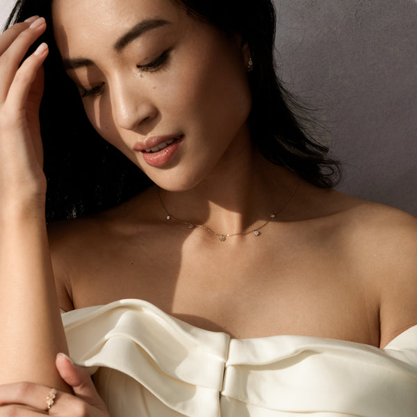 A close-up of a woman wearing a white ruffled off the shoulder top and delicate gold chain necklace with three dangling flowers and two dangling round clear stones, set against a dark grey background.