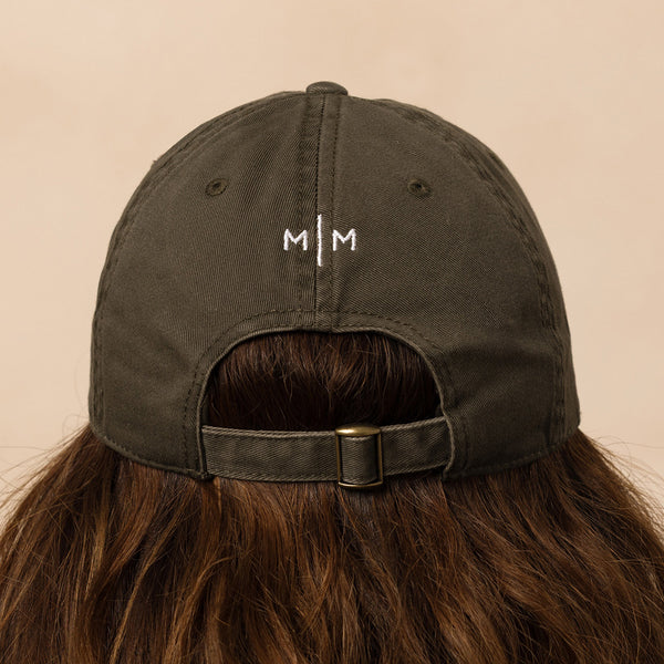 Dark green cap with 'M/M' logo worn by a person with brown hair on a beige background
