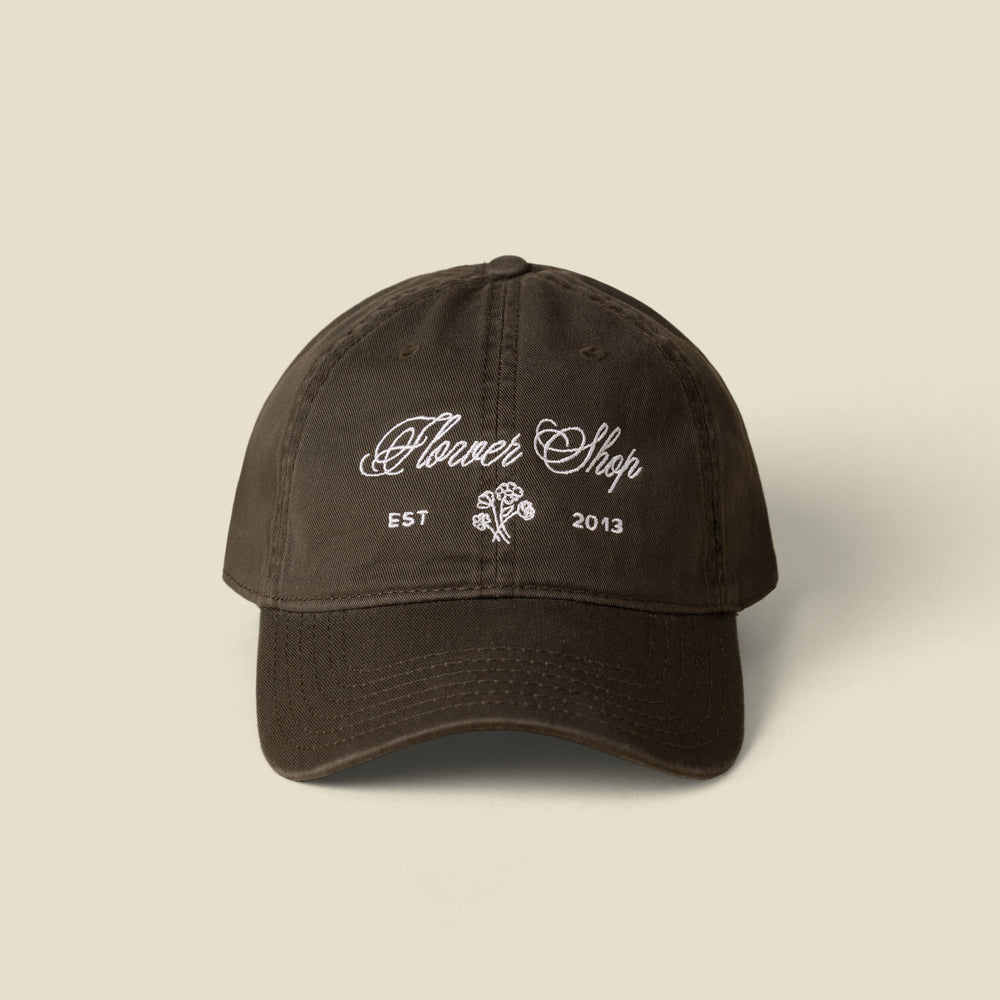 A dark green cotton baseball cap with white embroidered text reading 'Flower Shop' and 'EST 2013' with a small rose graphic on the front, shown against a solid light beige background.
