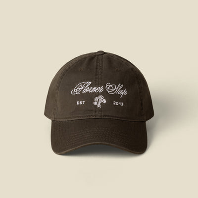 A dark green cotton baseball cap with white embroidered text reading 'Flower Shop' and 'EST 2013' with a small rose graphic on the front, shown against a solid light beige background.