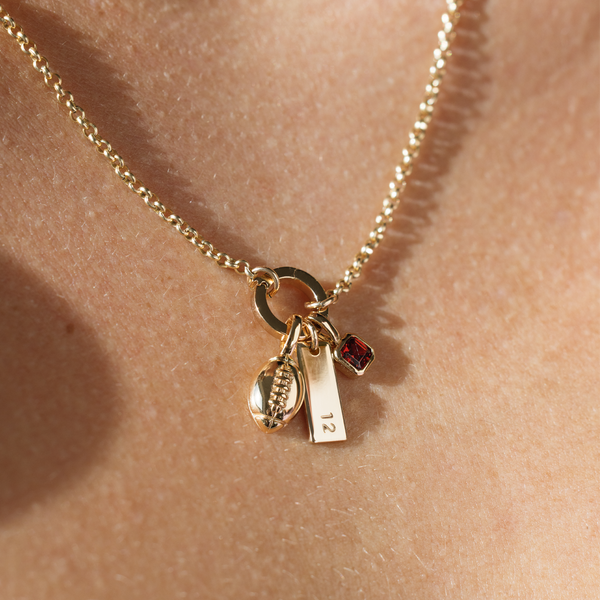 A close-up image of a person wearing a gold chain charm necklace with a round mechanism holding the charms, featuring a gold football charm, a small vertical bar charm engraved with the number '12', and a square, bezel-set red charm.