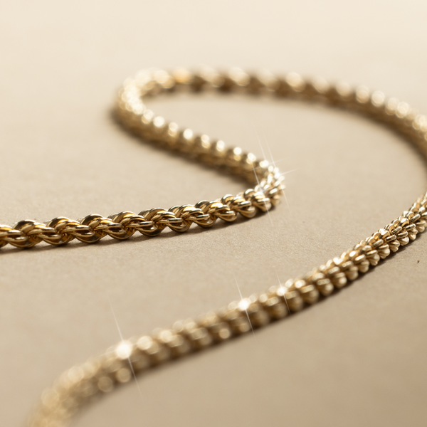 A close-up of a gold rope texture chain necklace, set on a neutral background.