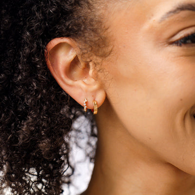 A close-up of a person's ear wearing two gold hoop earrings, one with clear gemstones in a curving pattern and the other with the word 'GEMINI' in raised letters across the front.