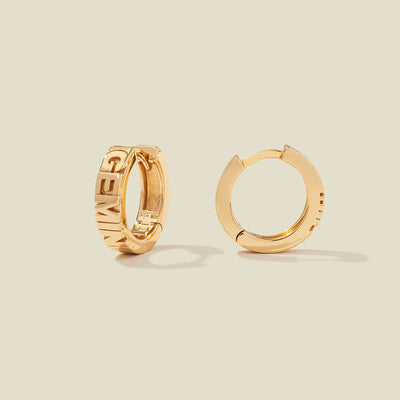 A pair of gold huggie hoop earrings on a light beige background. One earring is shown from the side, displaying a polished outer surface, while the other is shown from the front, showcasing the word 'GEMINI' in raised letters.