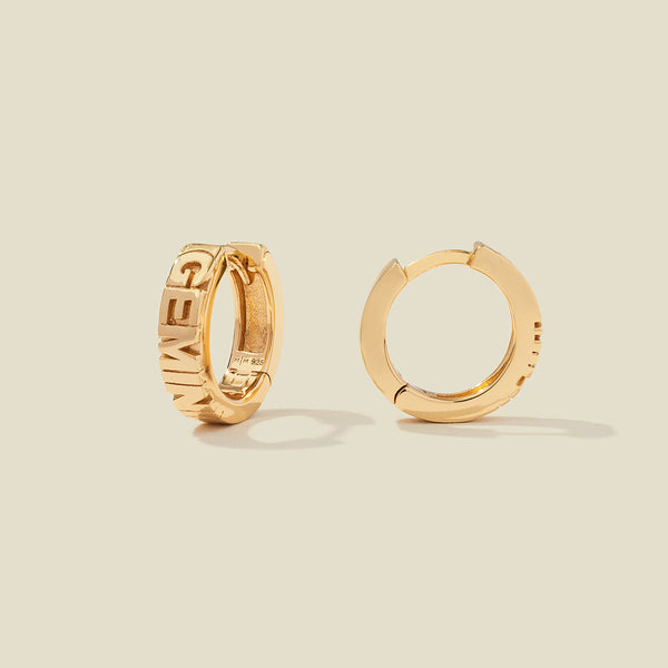 A pair of gold huggie hoop earrings on a light beige background. One earring is shown from the side, displaying a polished outer surface, while the other is shown from the front, showcasing the word 'GEMINI' in raised letters.