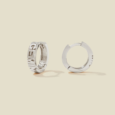 A pair of silver huggie hoop earrings on a light beige background. One earring is shown from the side, displaying a polished outer surface, while the other is shown from the front, showcasing the word 'GEMINI' in raised letters.