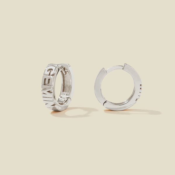 A pair of silver huggie hoop earrings on a light beige background. One earring is shown from the side, displaying a polished outer surface, while the other is shown from the front, showcasing the word 'GEMINI' in raised letters.