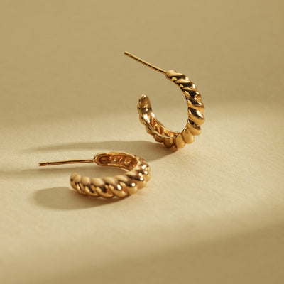 A pair of gold crescent-shaped hoop earrings with a textured, ridged outer surface, shown on a shadowed light beige background.