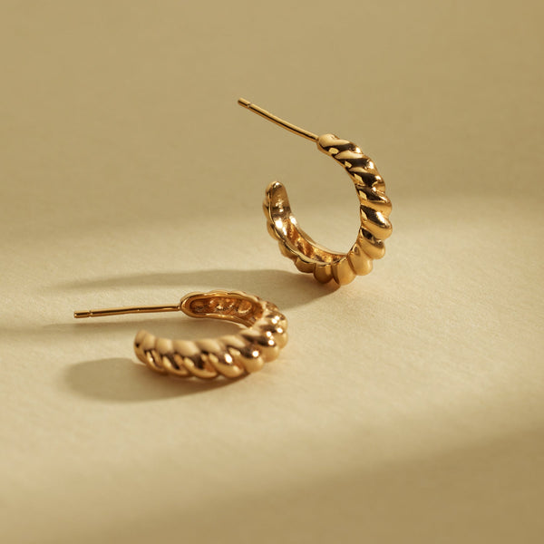 A pair of gold crescent-shaped hoop earrings with a textured, ridged outer surface, shown on a shadowed light beige background.