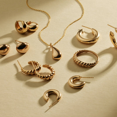 A variety of gold earrings, rings, and a necklace laying on a shadowed light beige surface.