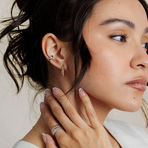 A person's ear wearing a ribbed texture gold hoop and a wide polished silver ear cuff, paired with gold and silver matching ribbed rings.