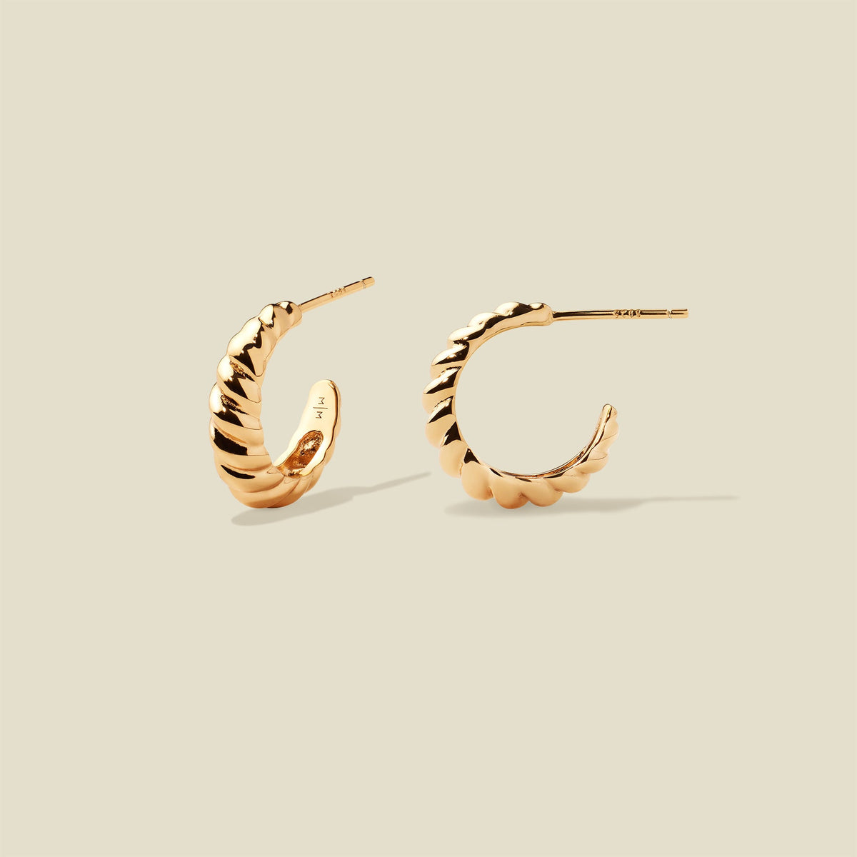A pair of gold crescent-shaped hoop earrings with a textured, ridged outer surface, shown on a solid light beige background.
