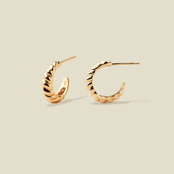 A pair of gold crescent-shaped hoop earrings with a textured, ridged outer surface, shown on a solid light beige background.