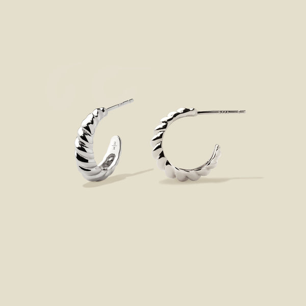 A pair of silver crescent-shaped hoop earrings with a textured, ridged outer surface, shown on a solid light beige background.