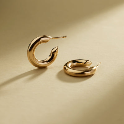 A pair of polished gold hoop earrings with a curved, open C-shape design, shown on a shadowed warm beige background.