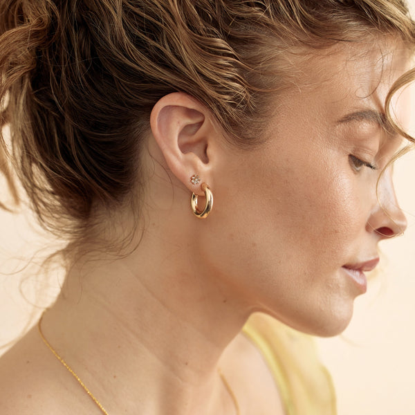 A close-up of a woman's ear wearing a polished gold hoop earring and a gold flower stud with clear stones.