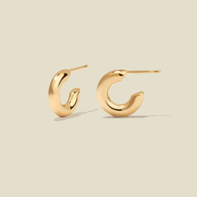 A pair of polished gold hoop earrings with a curved, open C-shape design, shown on a solid light beige background.