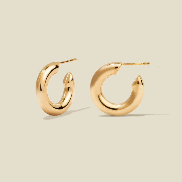 A pair of polished gold hoop earrings with a curved, open C-shape design, shown on a solid light beige background.