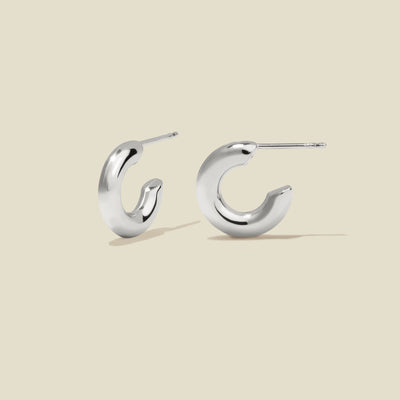 A pair of polished silver hoop earrings with a curved, open C-shape design, shown on a solid light beige background.