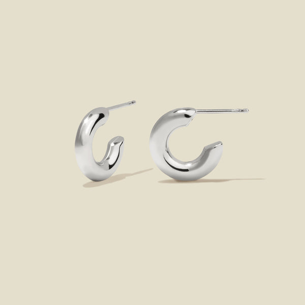 A pair of polished silver hoop earrings with a curved, open C-shape design, shown on a solid light beige background.