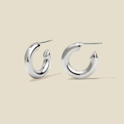 A pair of polished silver hoop earrings with a curved, open C-shape design, shown on a solid light beige background.