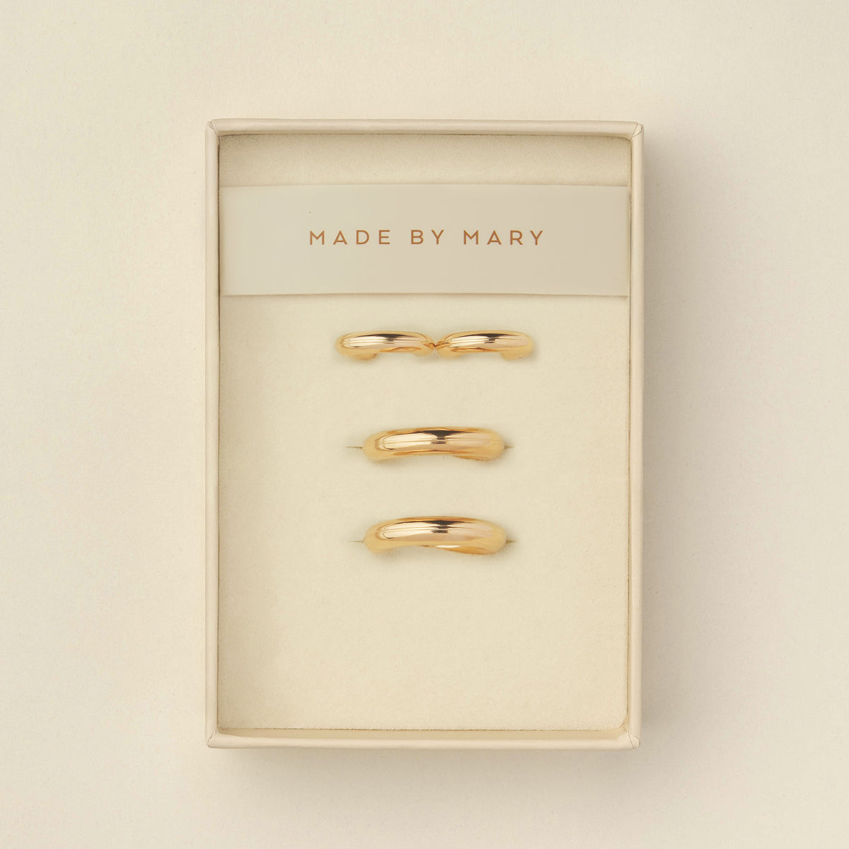 Two pairs of polished wide gold hoops, one smaller and one larger, displayed in a cream-colored presentation box with the brand name 'MADE BY MARY' printed on the insert.