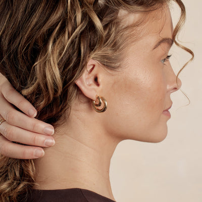 A close-up lifestyle image of a woman's ear wearing two pairs of wide polished, rounded gold hoops in different sizes.