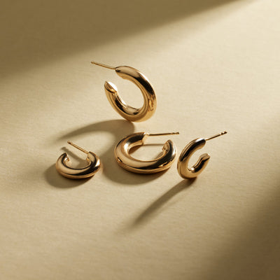 Two pairs of polished wide gold hoops in different sizes, some laying flat and others sitting upright on a textured beige background with natural shadows.