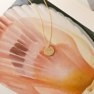 A delicate gold round pendant chain necklace with the word 'GRANDMA' engraved on the disc, shown on a watercolor painting of a shell with various pink, orange, and black hues.