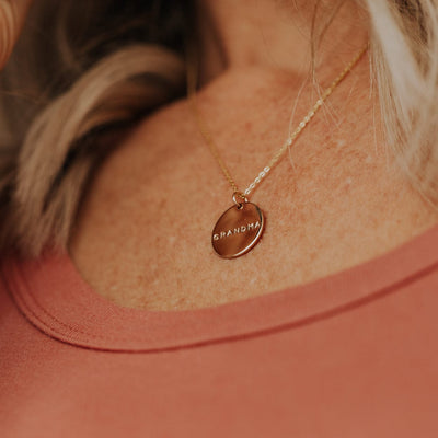A person wearing a delicate gold pendant chain necklace with a round flat disc engraved with the word 'GRANDMA'.
