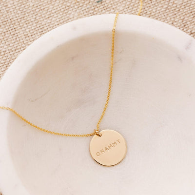 A delicate gold pendant chain necklace draped in a white marble bowl, with a round flat disc engraved with the word 'GRAMMY'.