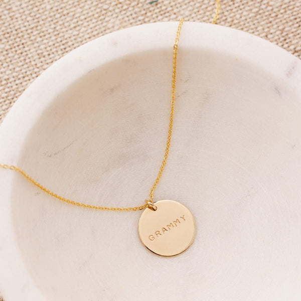 A delicate gold pendant chain necklace draped in a white marble bowl, with a round flat disc engraved with the word 'GRAMMY'.