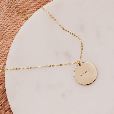 A delicate gold pendant chain necklace draped on a round white marble surface with a round flat disc charm engraved with the word 'MIMI', with a brown burlap texture in the background.