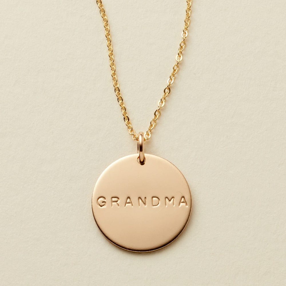 A gold chain necklace with a round gold disc pendant engraved with the word 'GRANDMA', displayed on a textured off-white background.