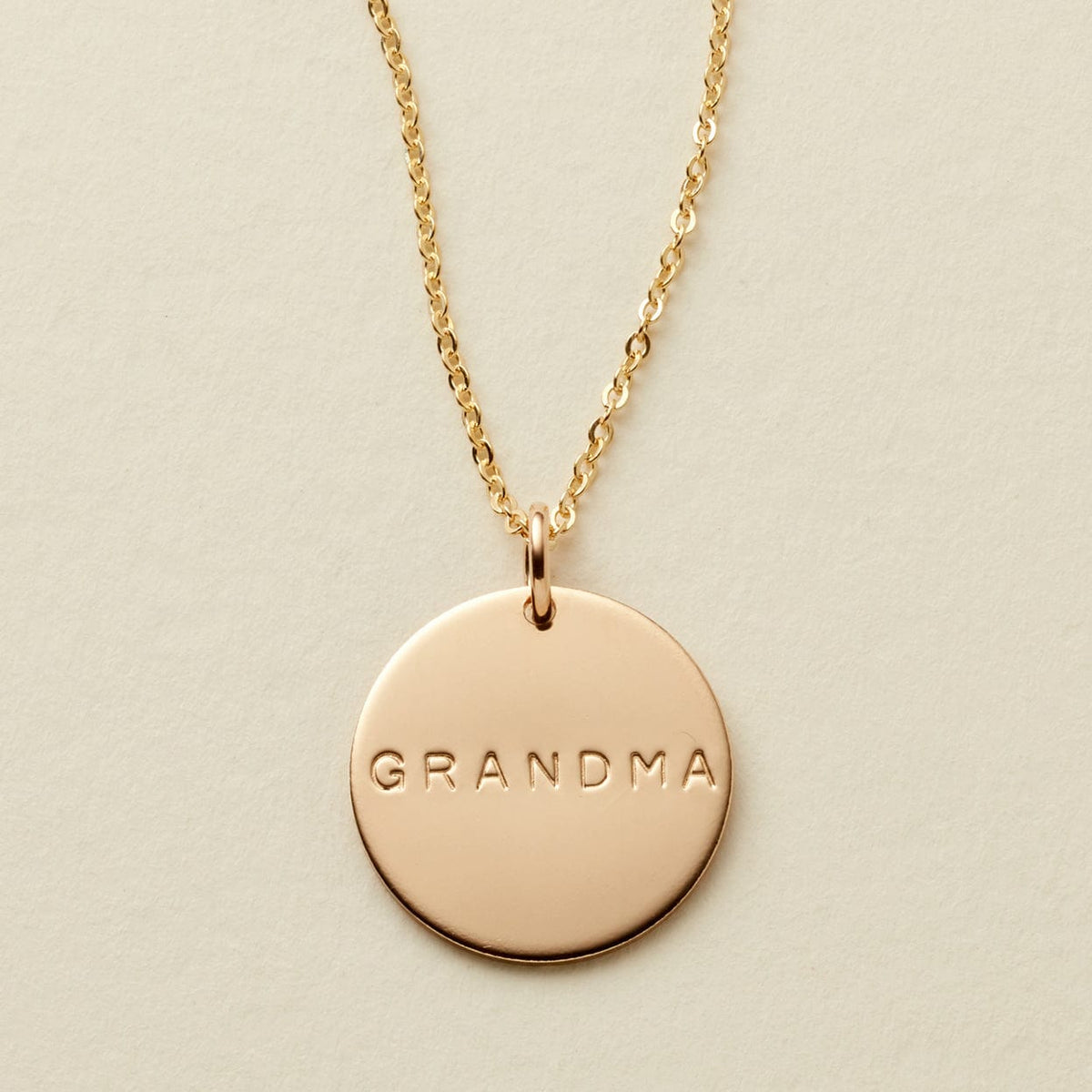 A gold chain necklace with a round gold disc pendant engraved with the word 'GRANDMA', displayed on a textured off-white background.