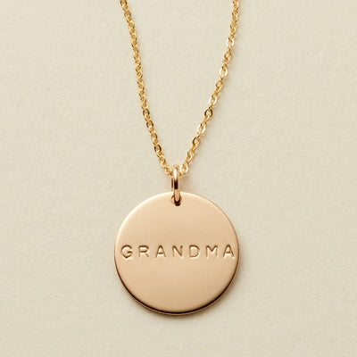 A gold chain necklace with a round gold disc pendant engraved with the word 'GRANDMA', displayed on a textured off-white background.
