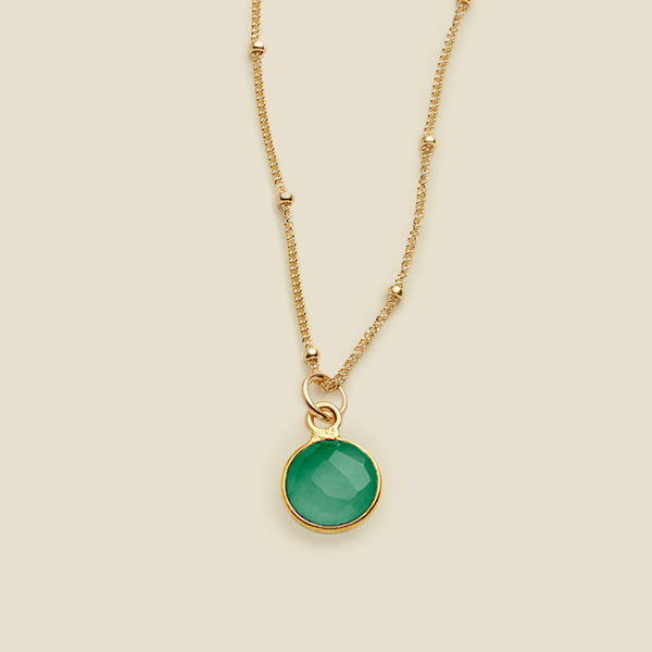 A gold chain pendant necklace with stationary gold beads, with an emerald green round gemstone charm, set against a solid beige background.