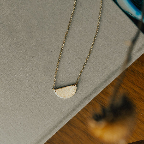 A gold chain necklace with a hammered half circle charm in the center, laying on a grey textured book on a dark wood surface.