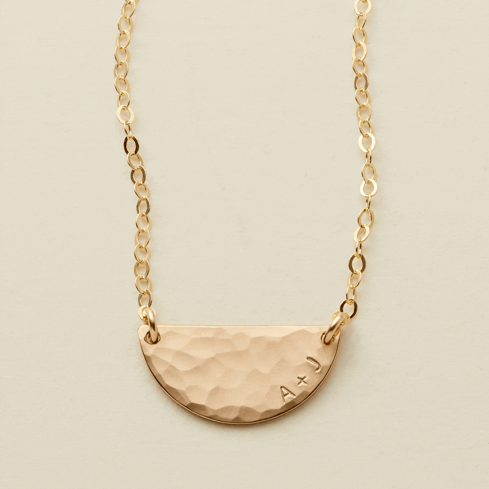 A gif of a delicate gold chain necklace with a hammered half circle charm centered on it, attached at both ends. It goes from being blank to having 'A + J' engraved on the right side along the curved edge.
