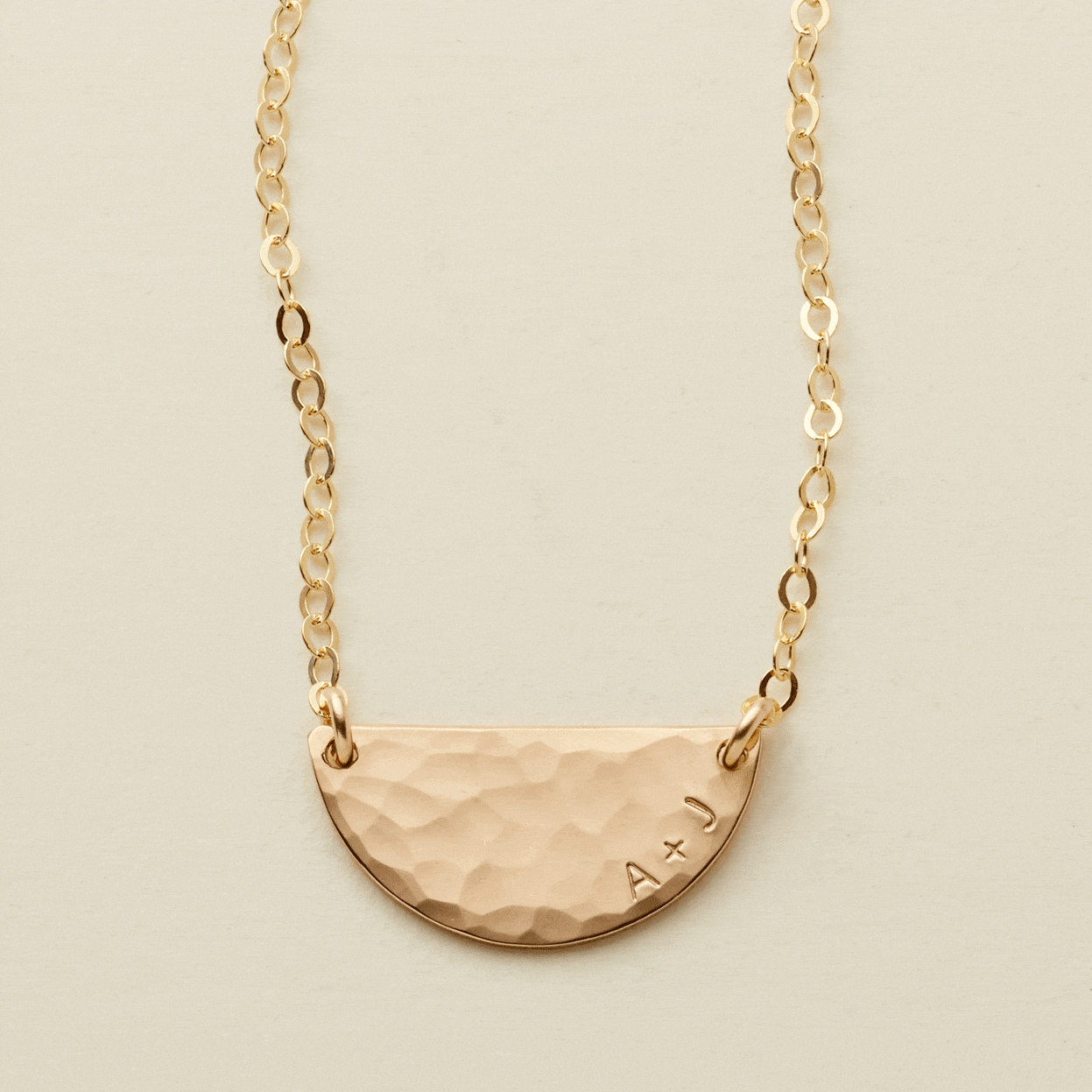 A gif of a delicate gold chain necklace with a hammered half circle charm centered on it, attached at both ends. It goes from being blank to having 'A + J' engraved on the right side along the curved edge.