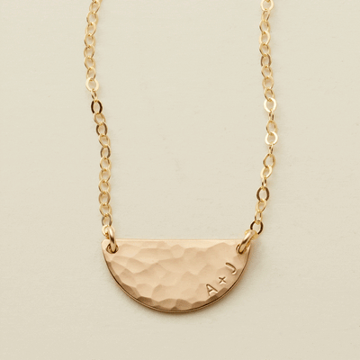A gif of a delicate gold chain necklace with a hammered half circle charm centered on it, attached at both ends. It goes from being blank to having 'A + J' engraved on the right side along the curved edge.