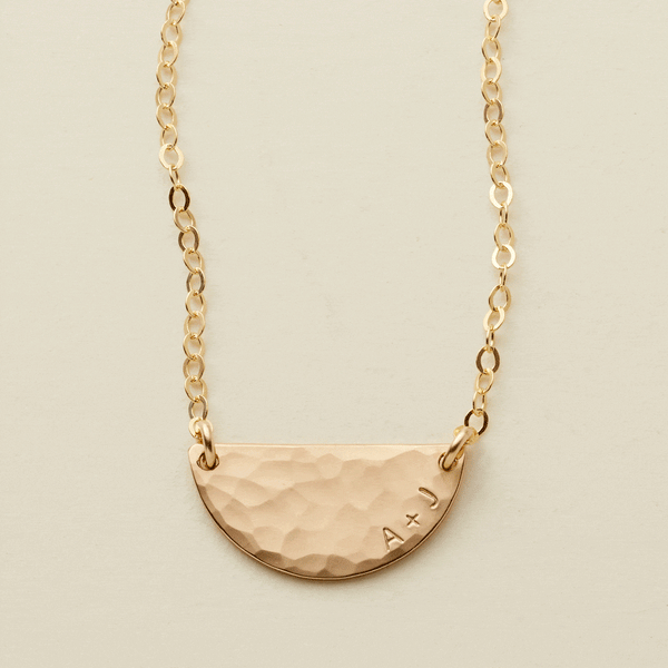 A gif of a delicate gold chain necklace with a hammered half circle charm centered on it, attached at both ends. It goes from being blank to having 'A + J' engraved on the right side along the curved edge.