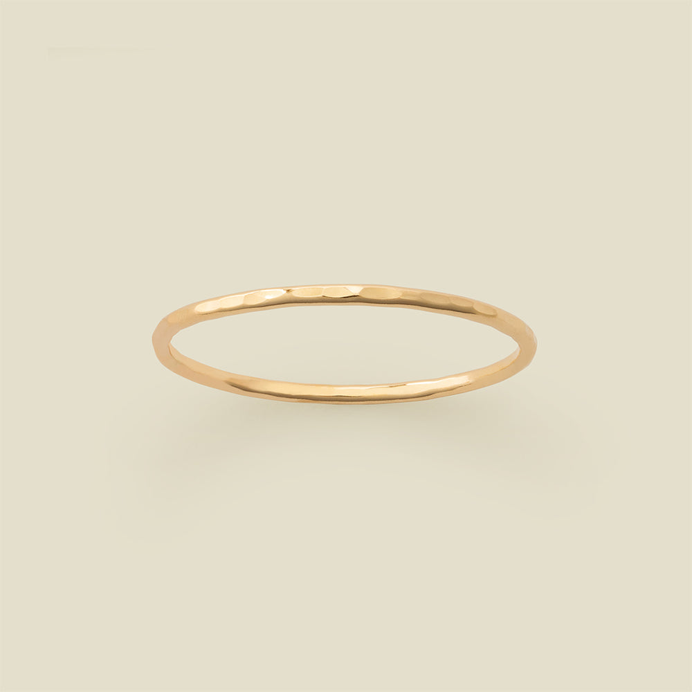 A thin hammered gold band ring centered on a solid light beige background.