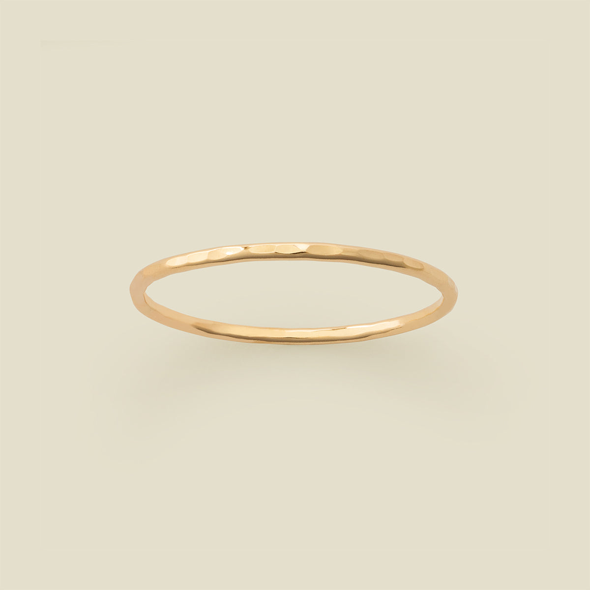 A thin hammered gold band ring centered on a solid light beige background.