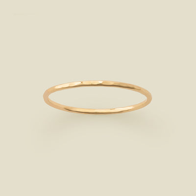 A thin hammered gold band ring centered on a solid light beige background.
