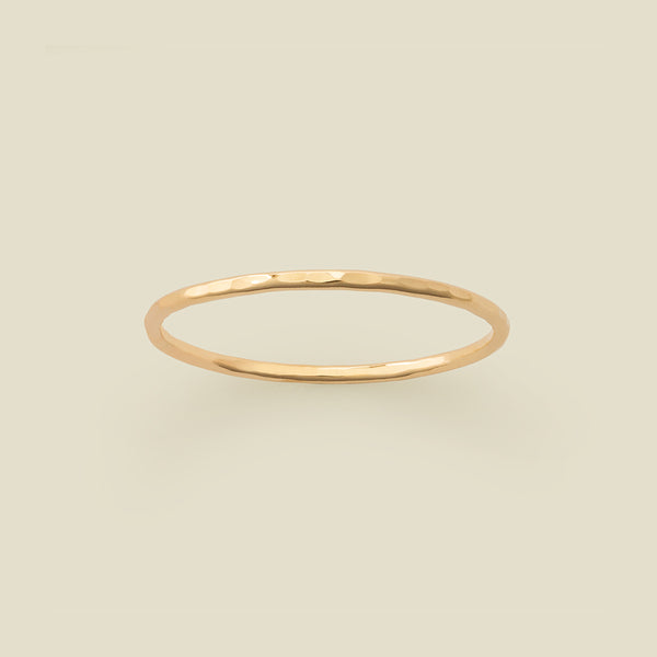 A thin hammered gold band ring centered on a solid light beige background.