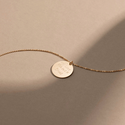 A gold chain necklace with a round gold pendant engraved with handwritten script, shown against a brown background.