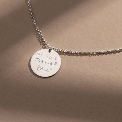 A silver chain necklace with a round silver pendant engraved with handwritten script, shown against a solid light brown background.