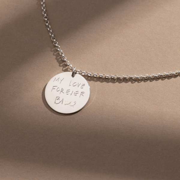 A silver chain necklace with a round silver pendant engraved with handwritten script, shown against a solid light brown background.