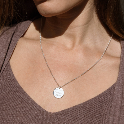 A lifestyle photo of a person's neck wearing a silver chain necklace with a round silver pendant engraved with handwritten script, along with a brown knit top.